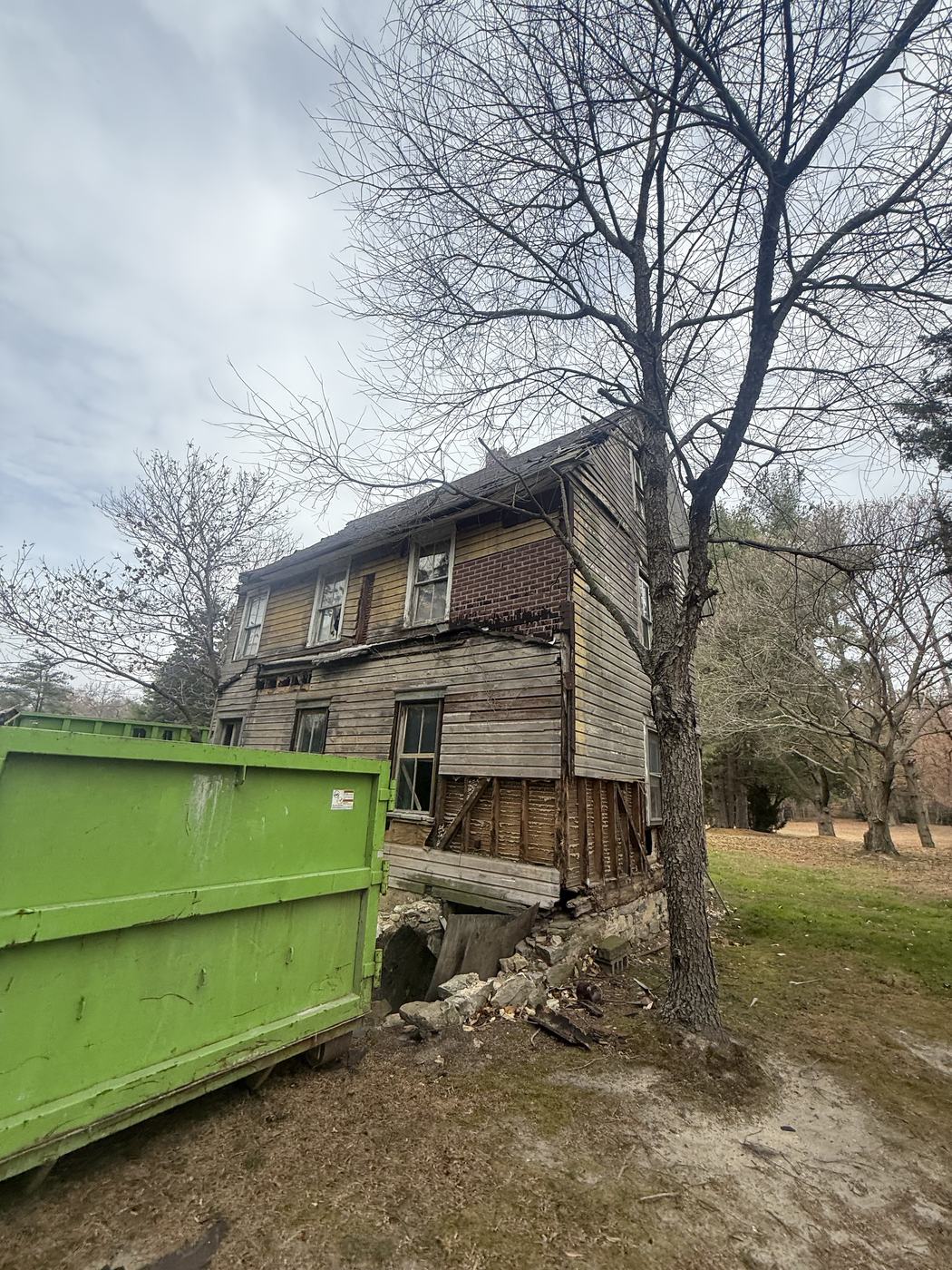 An old farmhouse beside a bare late fall tree with a roll off container staged alongside