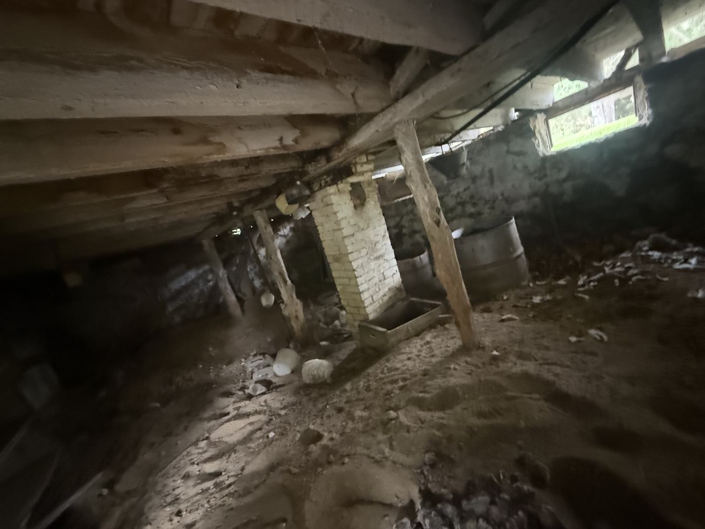 The crawl space under an old farmhouse showing hand cut timber joists, a masonry pier, and a dirt floor
