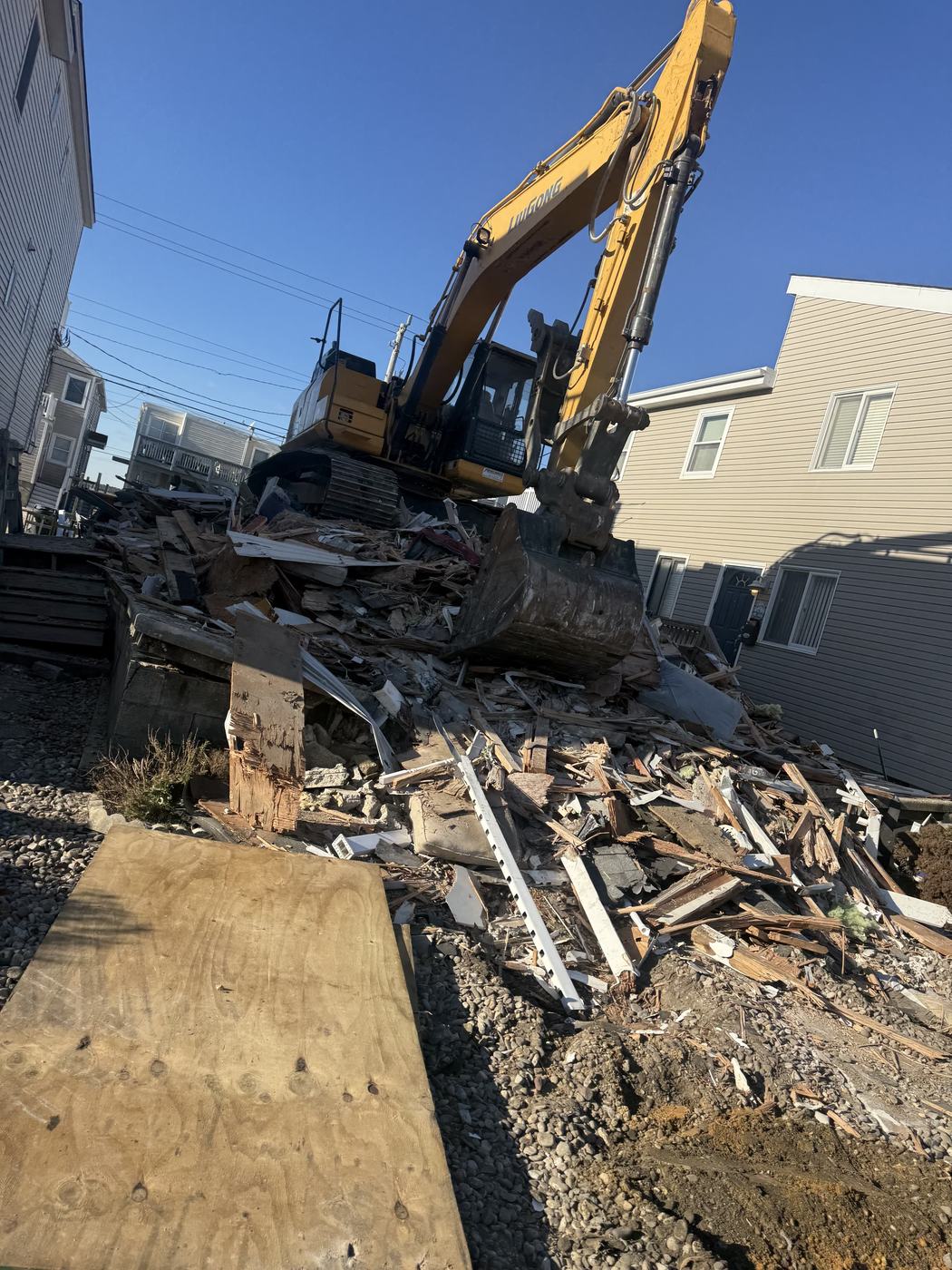 A debris pile staged on the home's own footprint with the excavator alongside