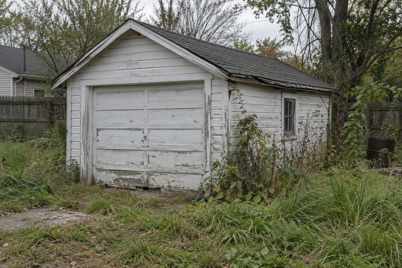A weathered detached single car garage with a sagging roofline in an overgrown backyard