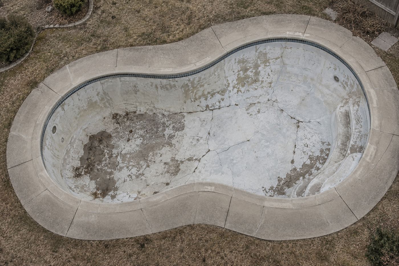 An empty cracked in ground swimming pool seen from above, drained and surrounded by concrete decking